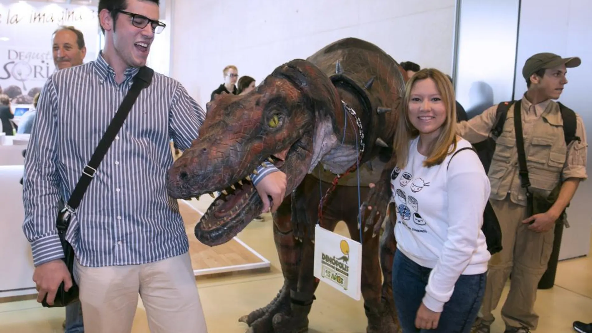Un dinosaurio promocionador del parque temático turolense de Dinópolis, bromea con unos visitantes dell XI Salón Aragonés del Turismo, Aratur, que se celebra en el Palacio de Congresos de la Expo de Zaragoza hasta el domingo y en el que tienen representación varias comunidades autónomas y algunos países, como Cuba
