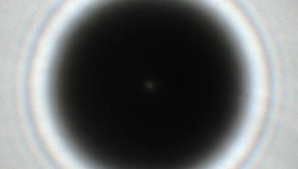 El punto de Arago (el punto claro en el centro del círculo negro) fue la primera demostración de que la luz es una onda. Lo que vemos en esta imagen es la sombra de un objeto circular colocado justo delante de un punto de luz. Intuitivamente pensaríamos que no puede haber luz en el interior de la sombra, y eso es lo que pasaría si la luz estuviera hecha de partículas. Pero una de las propiedades interesantes de las ondas es que pueden doblar ligeramente las esquinas, y en este caso eso significa que un poco de luz rodea el objeto y se cuela dentro de la sombra. Si, además, el punto luminoso coincide con el centro del objeto, esa luz “intrusa” se concentrará también en el centro, y eso es lo que vemos en la imagen. Por cierto, los círculos claros y oscuros alrededor de la sombra, y el borde borroso de ésta, también son consecuencia de las cosas que le pasan a la luz cerca del borde del objeto.
