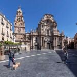 Dos personas pasean a sus perros en la Plaza del Cardenal Belluga, con la catedral de Murcia al fondo