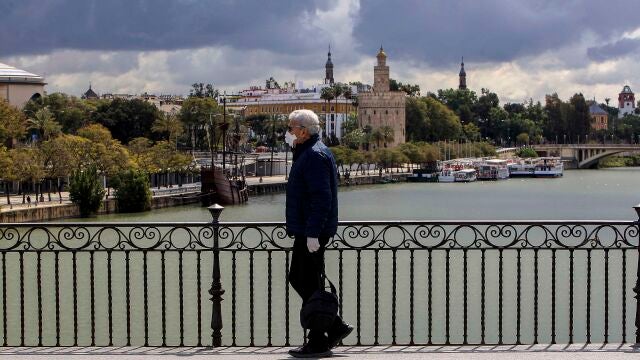 Un hombre pasea por el Puente de Triana de Sevilla con una mascarilla y guantes