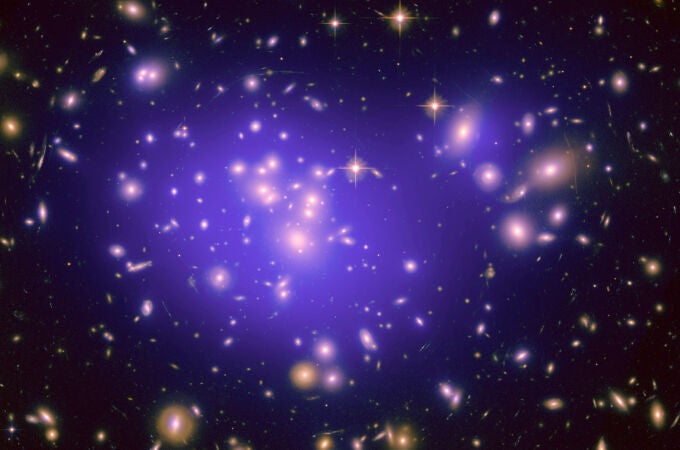 Aquí podemos ver el cúmulo de galaxias Abell 1689, con la nube de materia oscura en la que está bañado marcada en violeta. Observamos que la materia oscura se concentra en el centro del cúmulo, y también en las galaxias más masivas, como las que hay arriba a la izquierda y arriba a la derecha. La materia oscura no emite luz, así que esta nube la hemos reconstruido a partir de su gravedad, que produce un efecto llamado lente gravitatoria, que deforma las imágenes de galaxias que están detrás del cúmulo. Podemos ver muchas de estas imágenes deformadas formando arcos alrededor del cúmulo, como haría una lente circular muy potente.