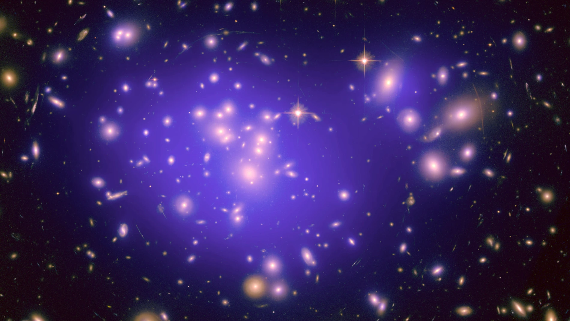 Aquí podemos ver el cúmulo de galaxias Abell 1689, con la nube de materia oscura en la que está bañado marcada en violeta. Observamos que la materia oscura se concentra en el centro del cúmulo, y también en las galaxias más masivas, como las que hay arriba a la izquierda y arriba a la derecha. La materia oscura no emite luz, así que esta nube la hemos reconstruido a partir de su gravedad, que produce un efecto llamado lente gravitatoria, que deforma las imágenes de galaxias que están detrás d...