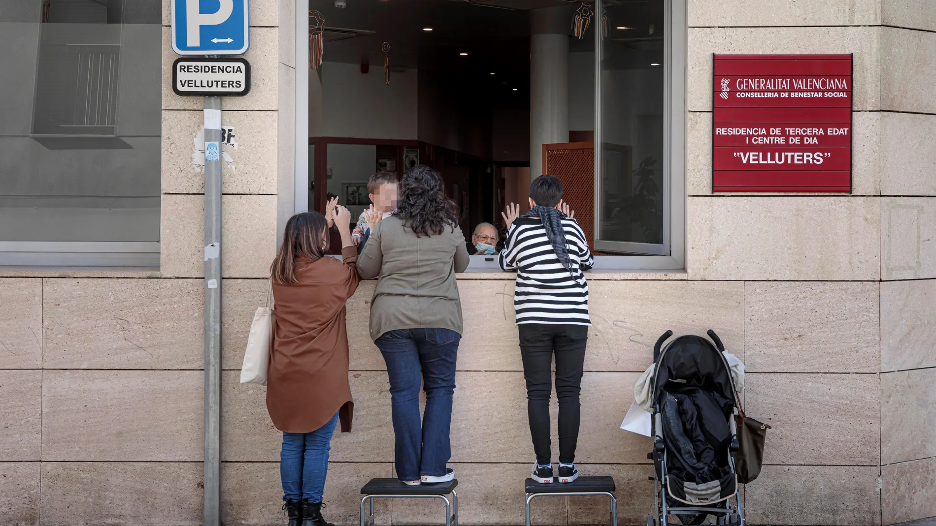 Una anciana recibe la visita de sus nietas a través de una ventana de la residencia de Velluters