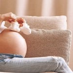 Pregnant woman holding small baby shoes relaxing at home on couch