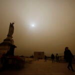 Varias personas caminan por el Puente Romano con la imagen del arcángel San Rafael de Córdoba con el cielo cubierto por la calima