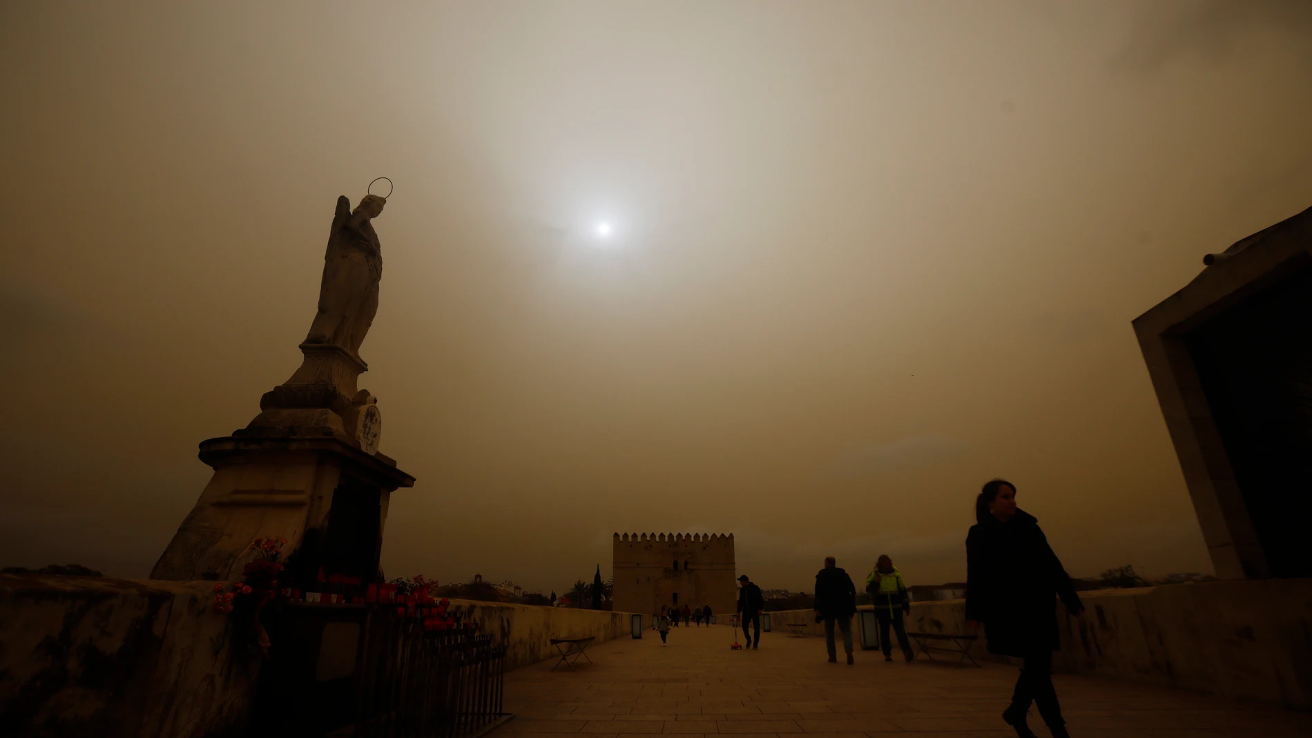 Varias personas caminan por el Puente Romano con la imagen del arcángel San Rafael de Córdoba con el cielo cubierto por la calima