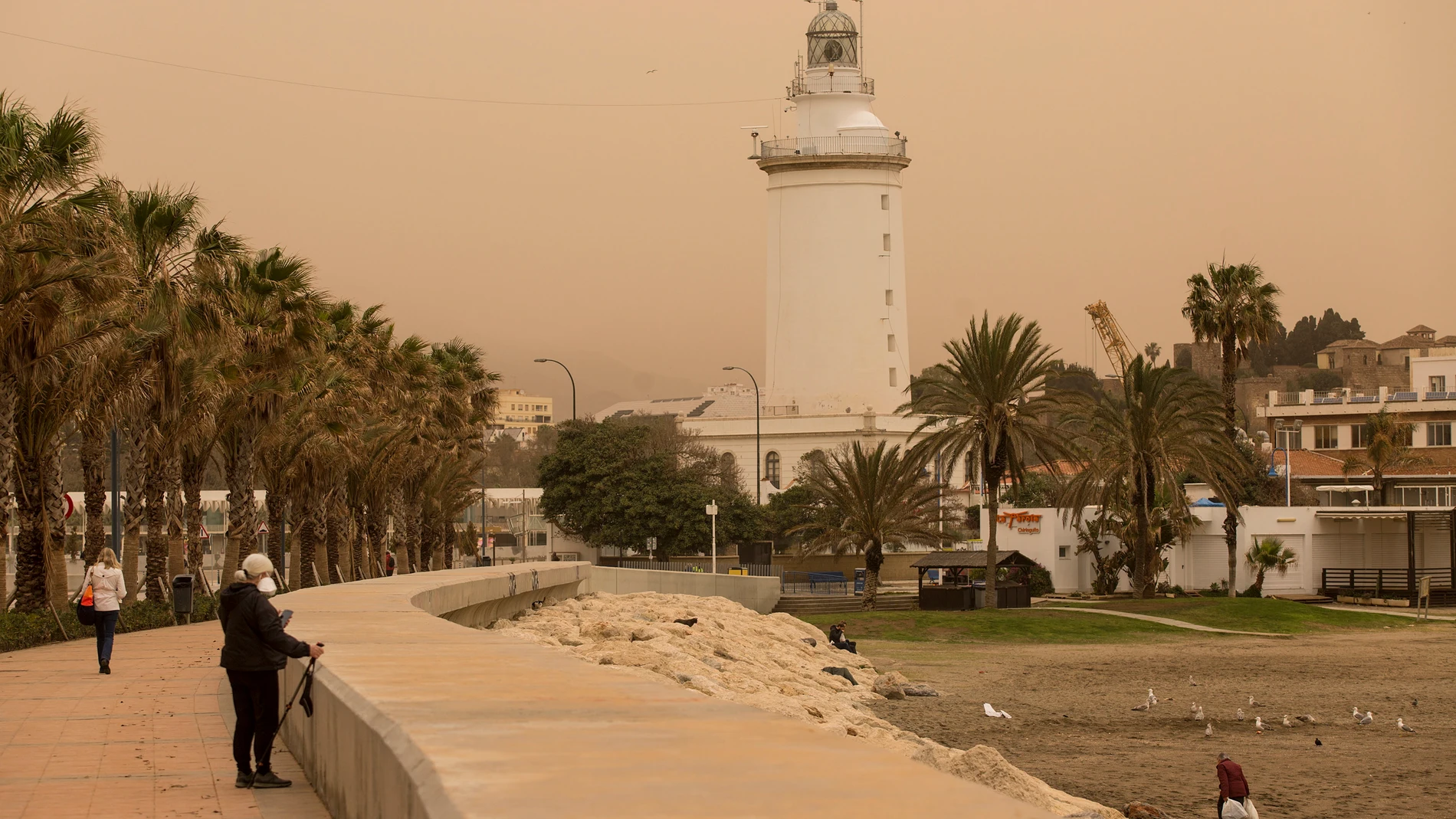 Varias personas caminan por la zona del Puerto de Málaga con La Farola al fondo que se encuentra cubierta por polvo procedente del desierto del Sáhara