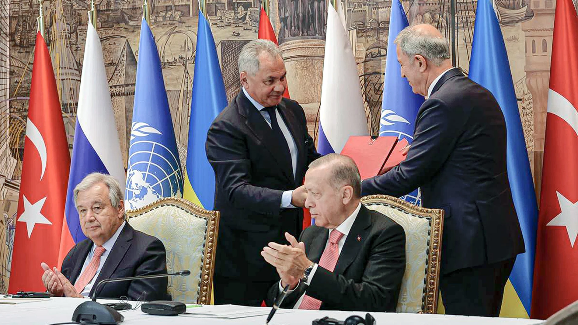 El presidente turco Recep Tayyip Erdogan, a la derecha, y el secretario general de la ONU, Antonio Guterres, se sientan mientras el ministro de Defensa ruso Sergei Shoigu, arriba a la izquierda, y el ministro de Defensa turco Hulusi Akar, arriba a la derecha, intercambian documentos durante una ceremonia de firma en el Palacio de Dolmabahce en Estambul, Turquía, el viernes 22 de julio de 2022.