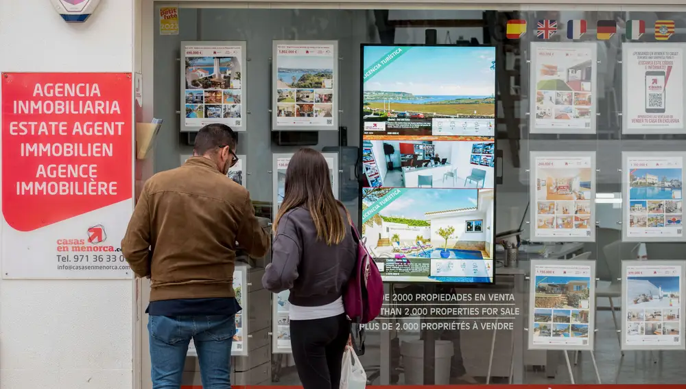 Una pareja se detiene a observar las propiedades en venta anunciadas en el escaparate de una agencia inmobiliaria de Menorca.