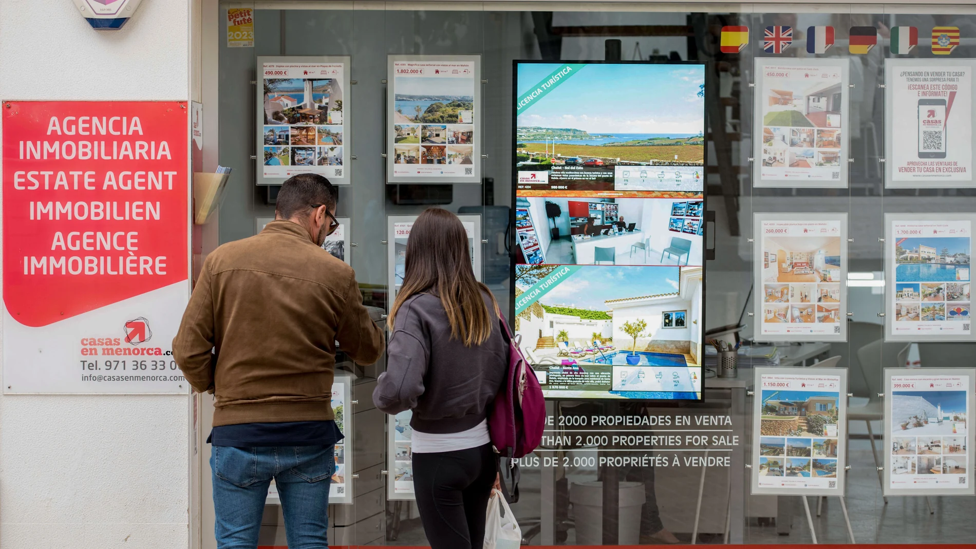 Una pareja se detiene a observar las propiedades en venta anunciadas en el escaparate de una agencia inmobiliaria de Menorca.