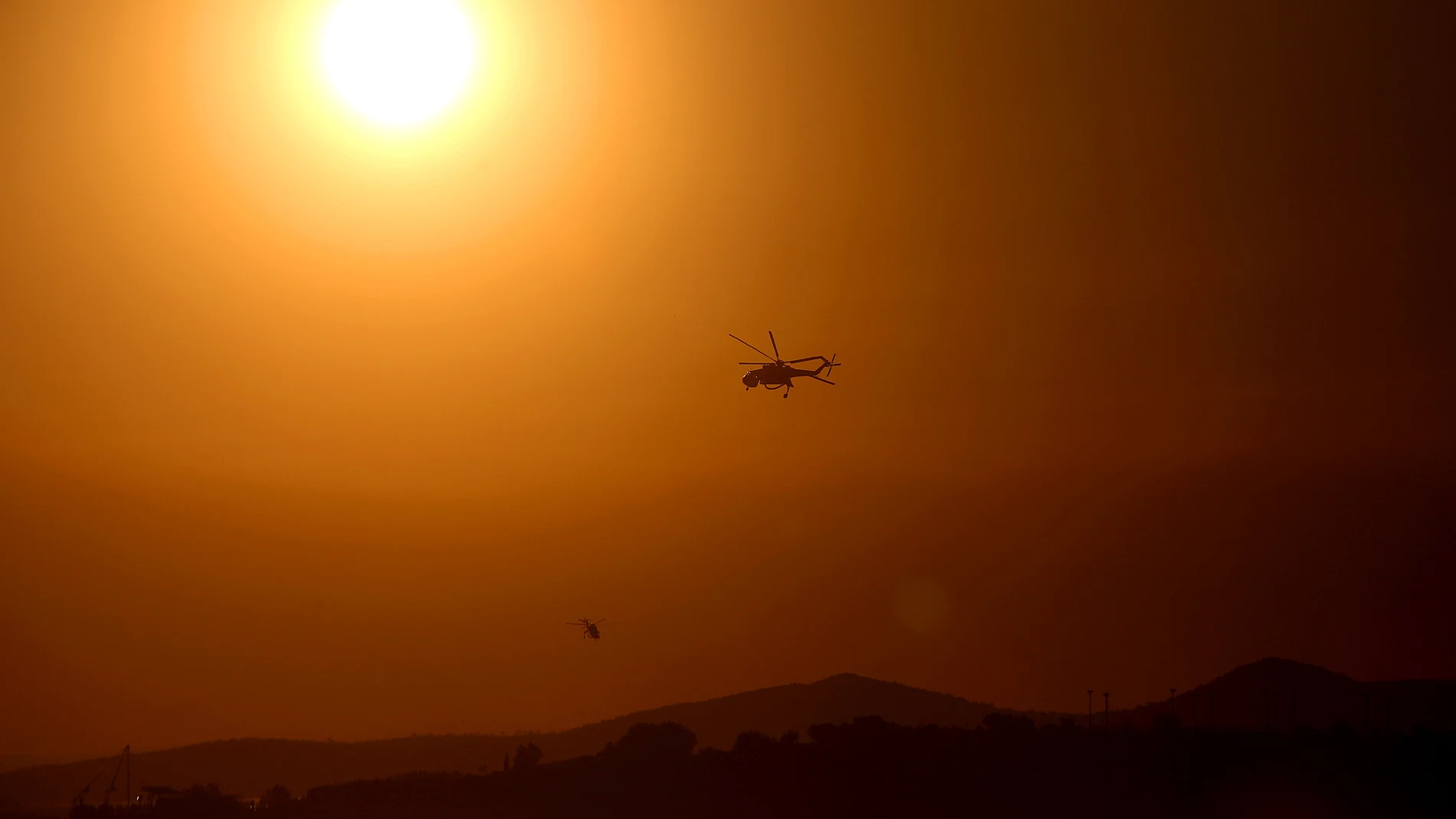 Un sol abrasador en Grecia mientras un helicóptero se dispone a cargar agua contra un incendio