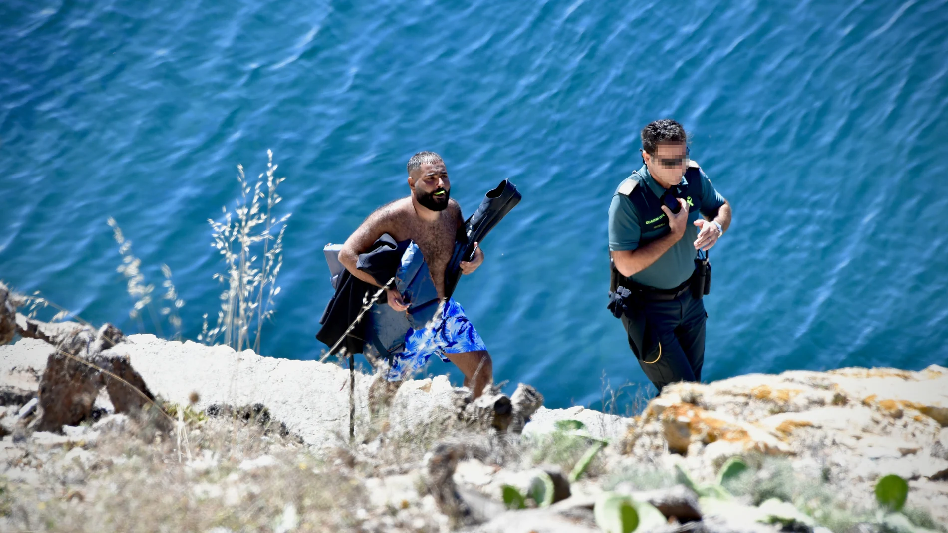 Un agente de la Guardia Civil con uno de los migrantes sirios que ha llegado a nado a las costas de Ceuta, en la barriada del Sarchal, a 31 de julio de 2023, en Ceuta (España). La Guardia Civil ha interceptado a dos migrantes sirios en una zona de rocas de la barriada del Sarchal de Ceuta. Los dos hombres venían equipados con trajes de neopreno y aletas y se prevé que hayan sido arrojados al mar a varias millas de la costa de Ceuta. Los agentes les han intervenido pasaportes sirios y los han trasladado a dependencias policiales en la frontera del Tarajal para la identificación y los trámites de filiación. 31 JULIO 2023;CEUTA;MIGRANTES;SIRIOS Antonio Sempere / Europa Press 31/07/2023