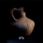 El pendiente de oro encontrado por los científicos en el Tossal de Baltarga, fotografiado sobre un fondo oscuro, frente a la vasija en la que fue hallado.