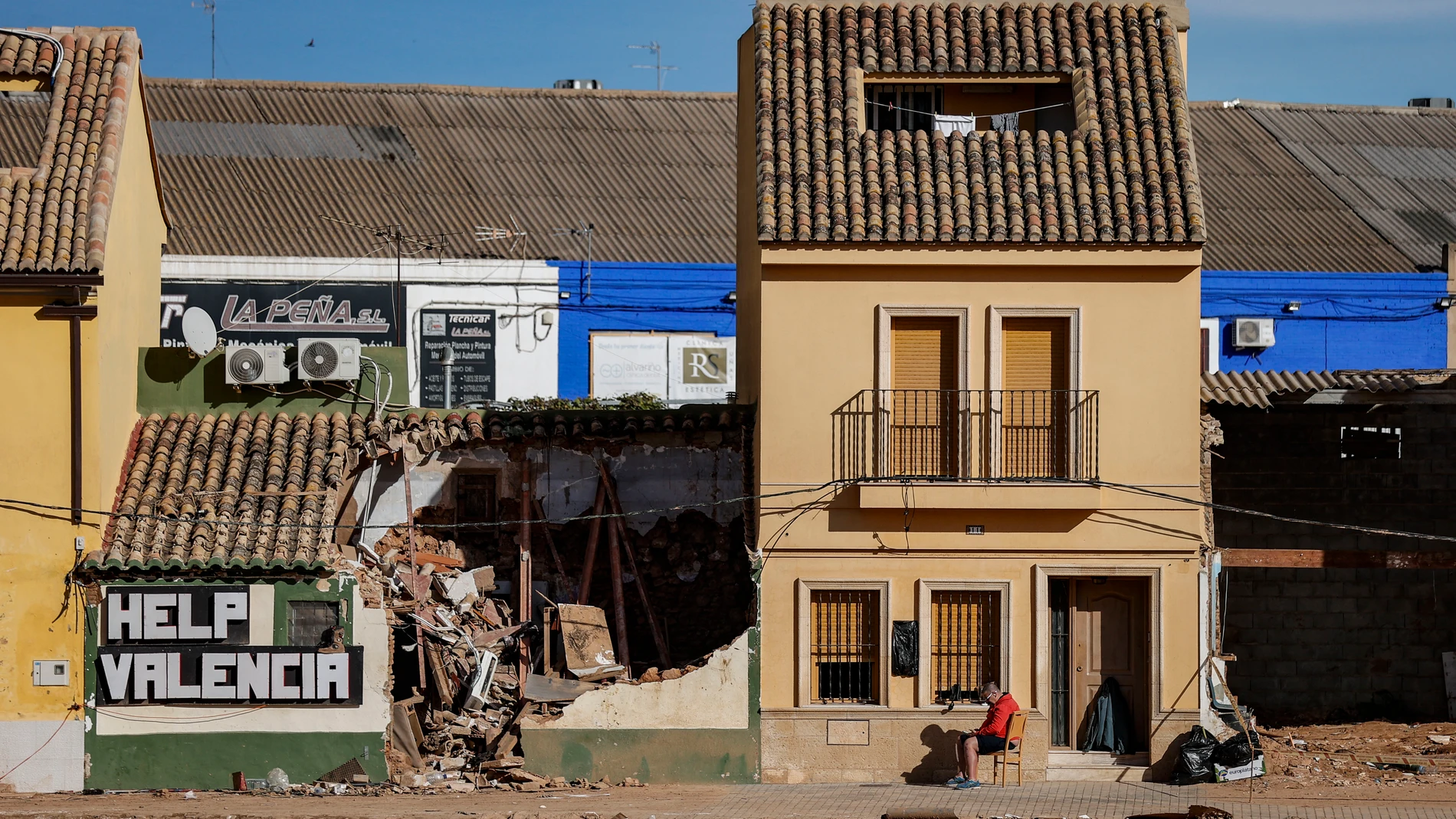 GRAFCVA4175. PICANYA (VALENCIA), 06/12/2024.- Un hombre consulta su teléfono sentado a la puerta de su casa junto al barranco del Poyo en Picanya (Valencia), mientras la vivienda del costado, se ha venido abajo debido a la fuerza del agua en la inundación del pasado 29 de noviembre, y donde alguien ha dibujado un cartel de "Help Valencia" (Ayudad a Valencia). EFE/Manuel Bruque