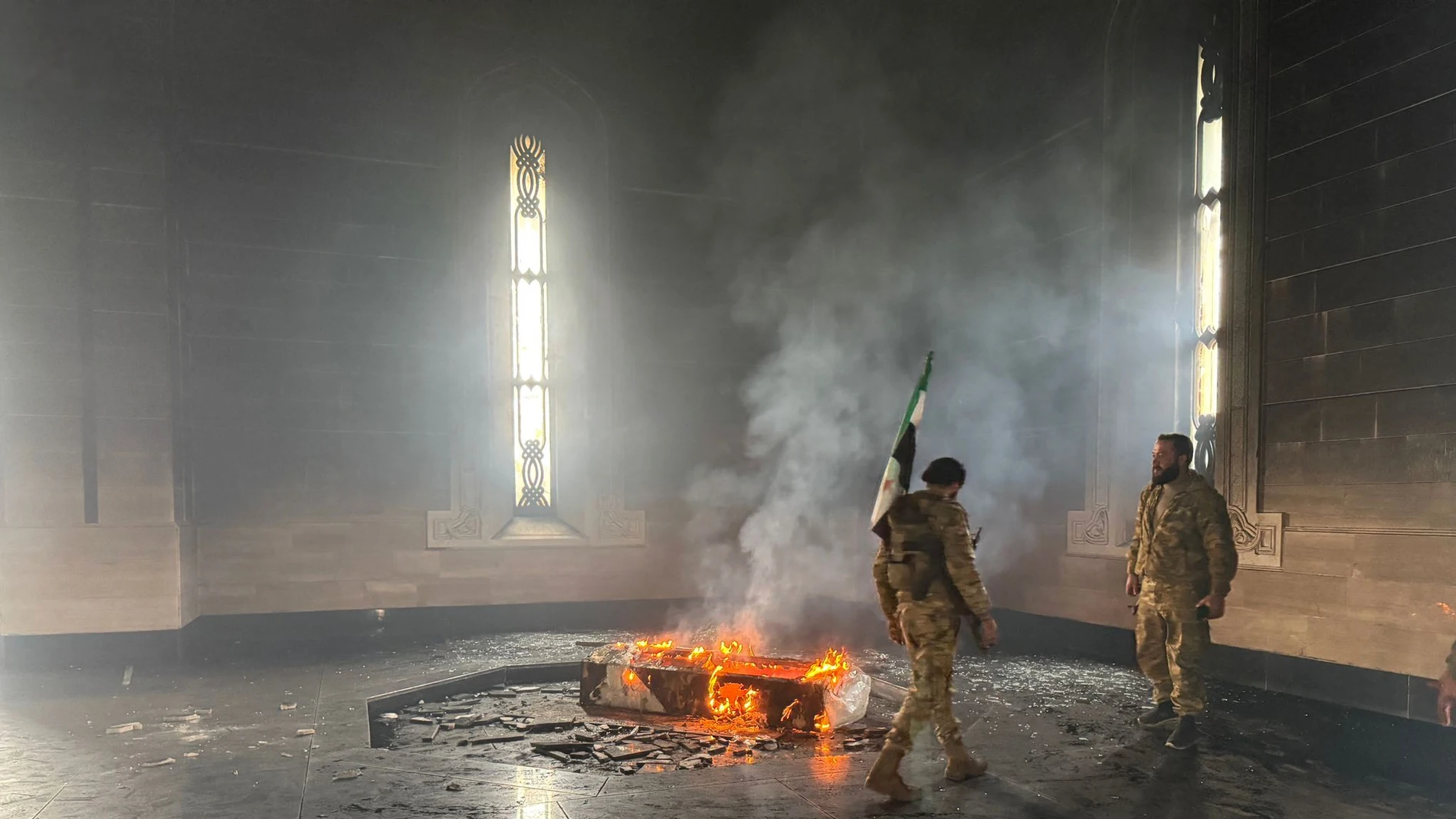 Rebel fighters stand next to the burning gravesite of Syria's late president Hafez al-Assad at his mausoleum in the family's ancestral village of Qardaha in the western Latakia province on December 11, 2024, after it was stormed by opposition factions. - The father and predecessor of Syria's ousted president Bashar al-Assad died in 2000.