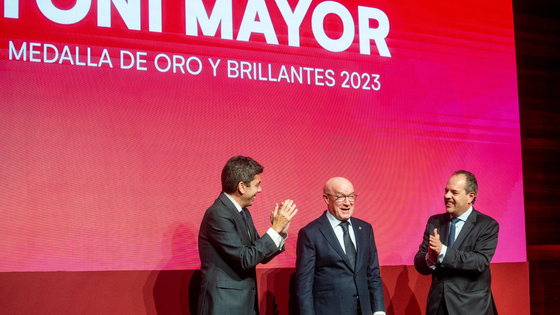 El presidente de la Generalitat Valenciana, Carlos Mazón, y el presidente de la Cámara de Comercio, Carlos Baño, entregan la medalla de oro y brillantes al expresidente de Hosbe, Toni Mayor