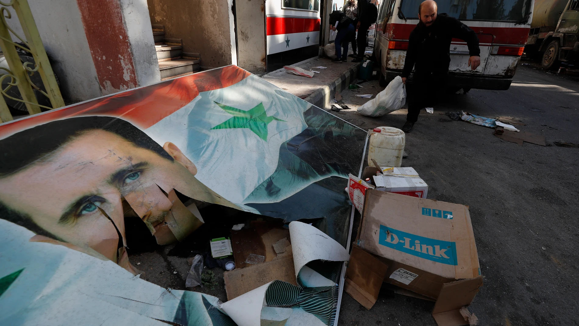 A Syrian member of the rebel forces prepares to destroy drugs and alcoholic beverages, found at a complex building, as he walks past damaged banner depicting the ousted Syrian President Bashar Assad, in Damascus, Syria, Wednesday, Dec. 25, 2024. (AP Photo/Omar Sanadiki)