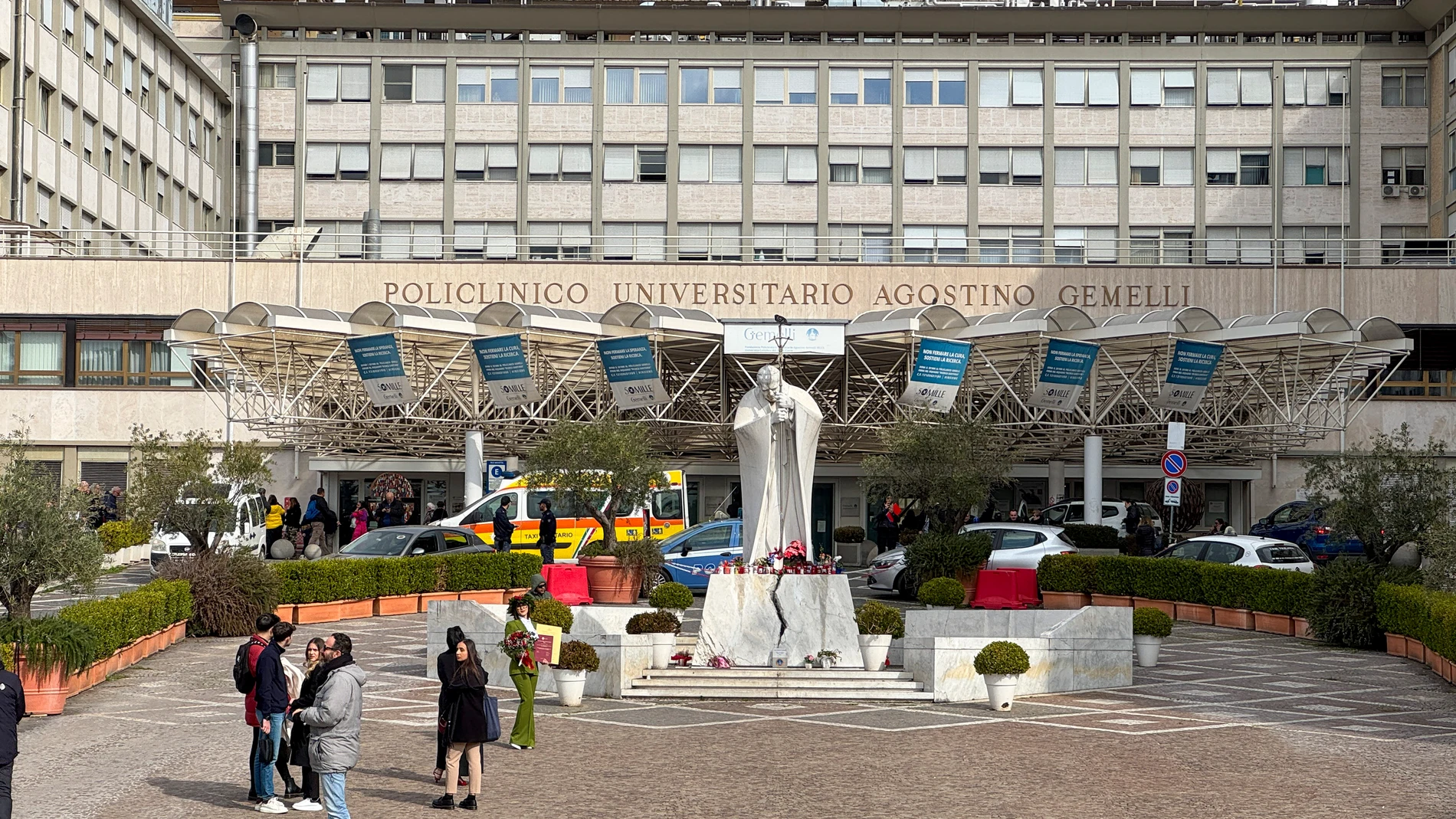 CIUDAD DEL VATICANO, 19/02/2025.- Velas y flores a los pies de la escultura de Juan Pablo II a las puertas del Hospital Gemelli de Roma donde permanece ingresado el papa Francisco, este miércoles. El papa pasó la noche tranquilo y desayunó en el sexto día de hospitalización en el Gemelli de Roma, informó el Vaticano, después de que este martes se comunicase un empeoramiento de su cuadro clínico al diagnosticarle una neumonía bilateral junto a la infección polimicrobiana que le obligó a ser in...