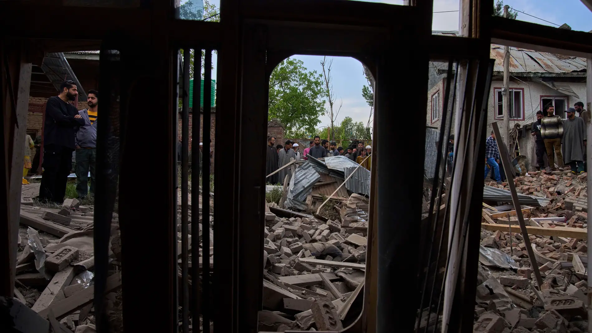 Kashmiri villagers inspect the debris of the blown-up family home of Ahsan Ul Haq Shiekh, a militant who officials say is involved in the deadly attack on tourists in Pahalgam, seen through the window of partially damaged house of a neighbor at Murran village in Pulwama, south of Srinagar, Indian controlled Kashmir, Saturday, April 26, 2025. (AP Photo/Dar Yasin)