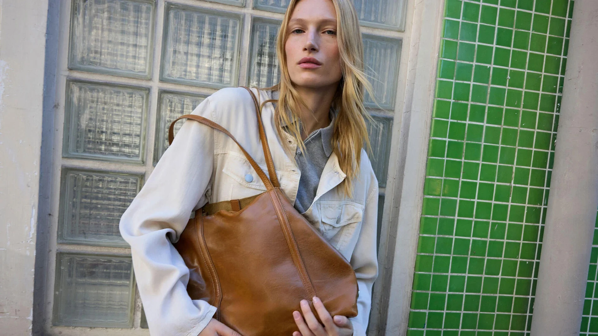 Bolso shopper marrón claro de Parfois llevado al hombro con chaqueta clara y fondo de azulejos verdes.