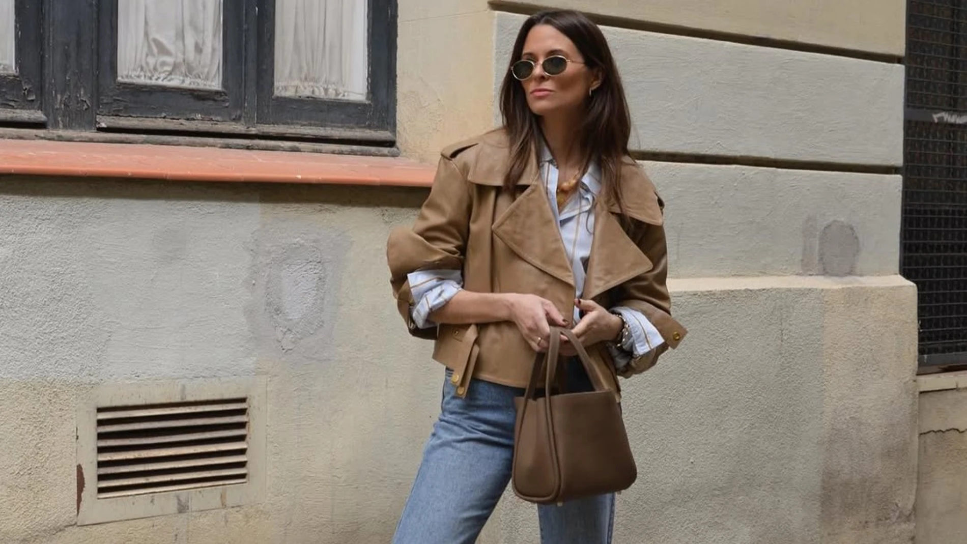 nfluencer con chaqueta corta en color neutro, camisa de rayas, vaqueros y bolso marrón, un conjunto en tendencia para el otoño 2025.