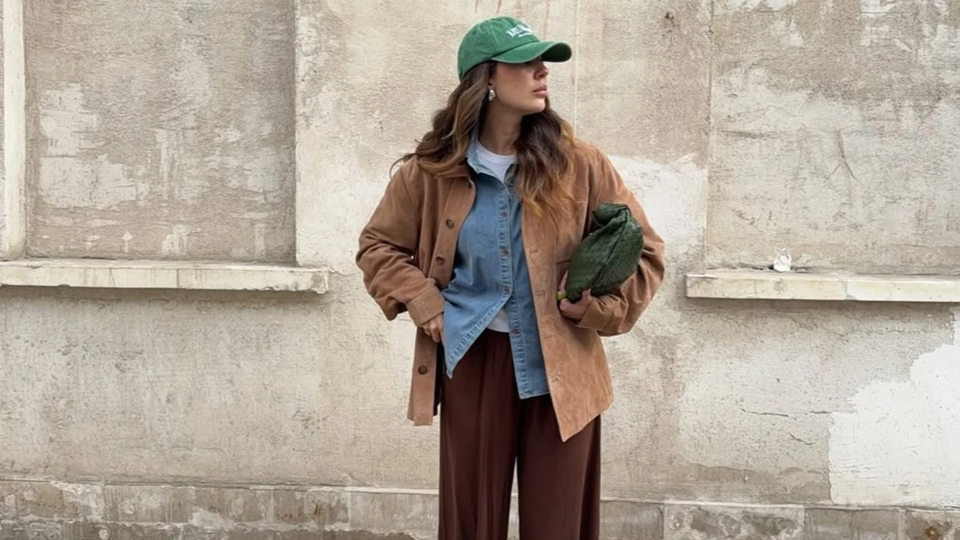 Mujer con chaqueta de ante marrón combinada con camisa denim, pantalones bombacho marrones y gorra verde, look de entretiempo.