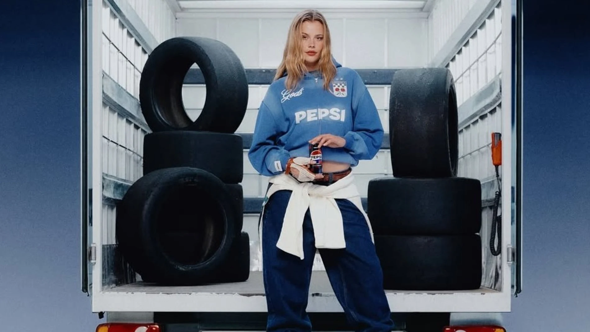 Modelo con sudadera azul de la colección Pepsi x Gods Brand, rodeada de neumáticos en un camión, imagen que refleja la inspiración racing de la cápsula.