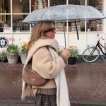 Influencer caminando bajo la lluvia con un paraguas transparente y un look otoñal en tonos beige.