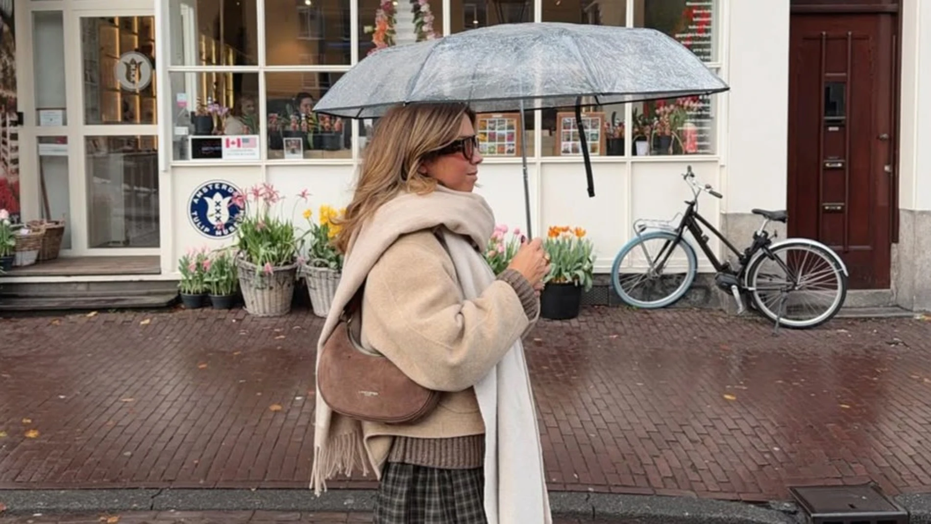 Influencer con abrigo de lana y bolso de ante, caminando bajo la lluvia con un paraguas transparente.