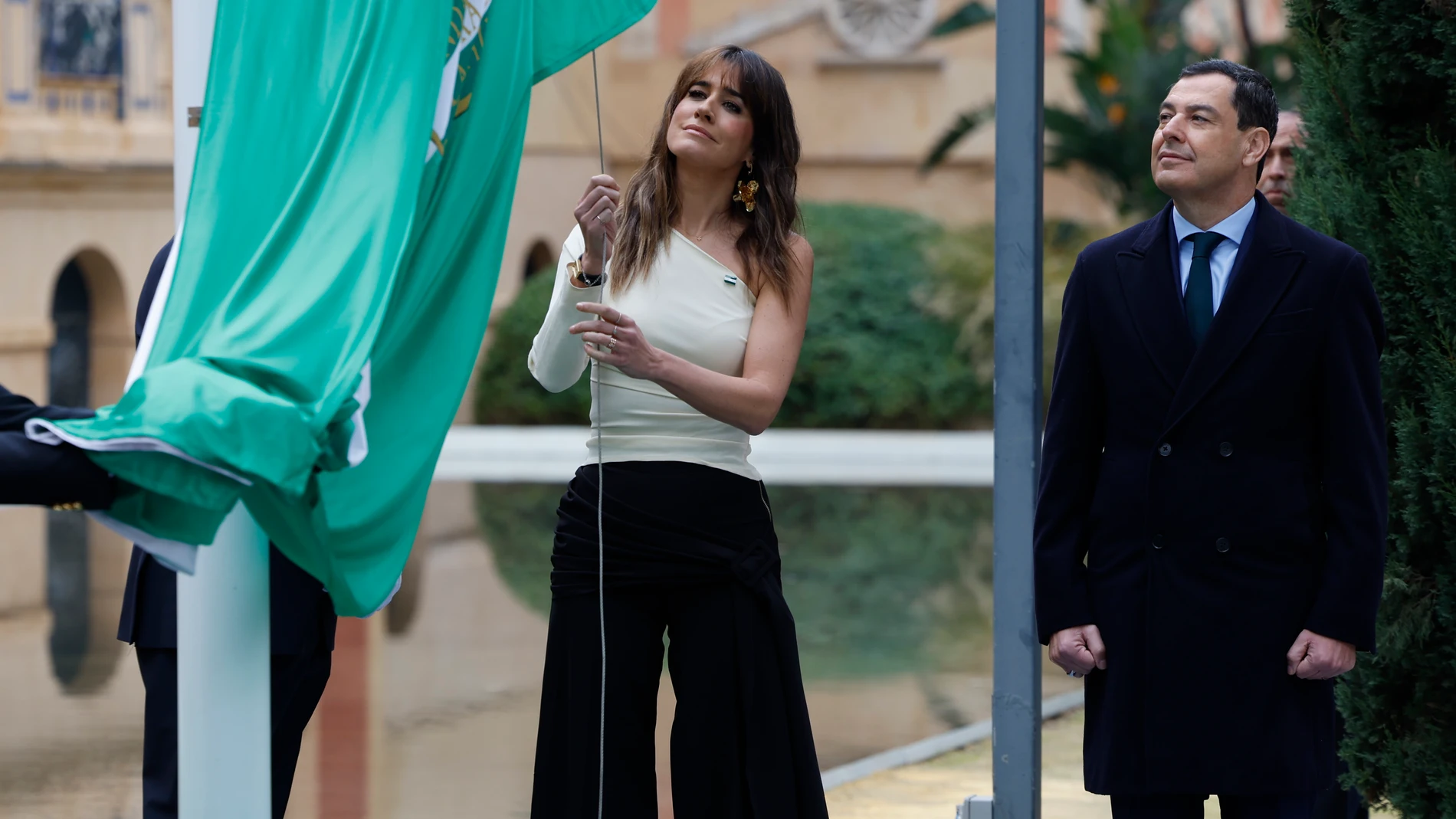 GRAFAND2443. SEVILLA, 04/12/2025.-El presidente andaluz, Juanma Moreno (d), observa el izado de la bandera de Andalucía por parte de la periodista Isabel Jiménez (c), autora este año del Elogio a la Bandera, en el mástil en los jardines del Palacio de San Telmo, sede de la Presidencia andaluza, durante el acto institucional del Día de la Bandera, que conmemora el aniversario de las multitudinarias manifestaciones del 4 de diciembre de 1977 para reclamar la autonomía andaluza, este jueves en S...