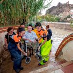 Voluntarios de protección civil trasladan a un hombre en silla de ruedas tras las intensas lluvias esta tarde en el barrio de Runes, Blanca