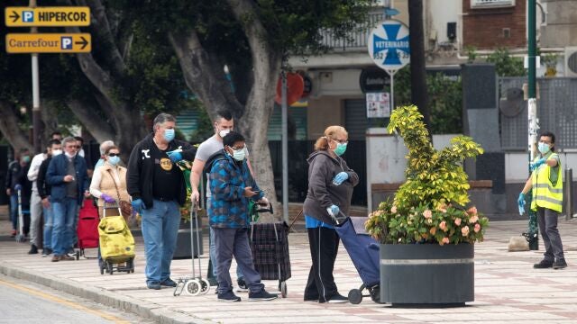 Varias personas esperan en una cola para entrar a un supermercado protegiéndose con mascarillas y guantes.