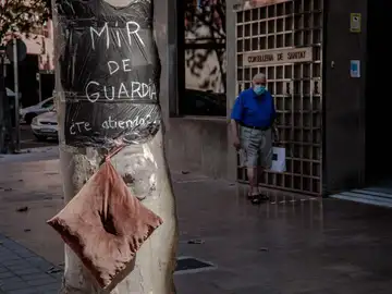 Una persona pasa ante la Consellería de Sanidad donde se puede ver una bolsa de basura usada a modo de bata con el lema "MIR de guardía ¿Te atiendo?" colgada de un arból, donde un grupo de médicos internos residentes (MIR) ha pasado esta noche al raso en el marco de la huelga indefinida que este colectivo mantiene para reivindicar mejores condiciones laborales. Los MIR pasan la noche al raso ante Sanidad y seguirán hasta que les atiendan