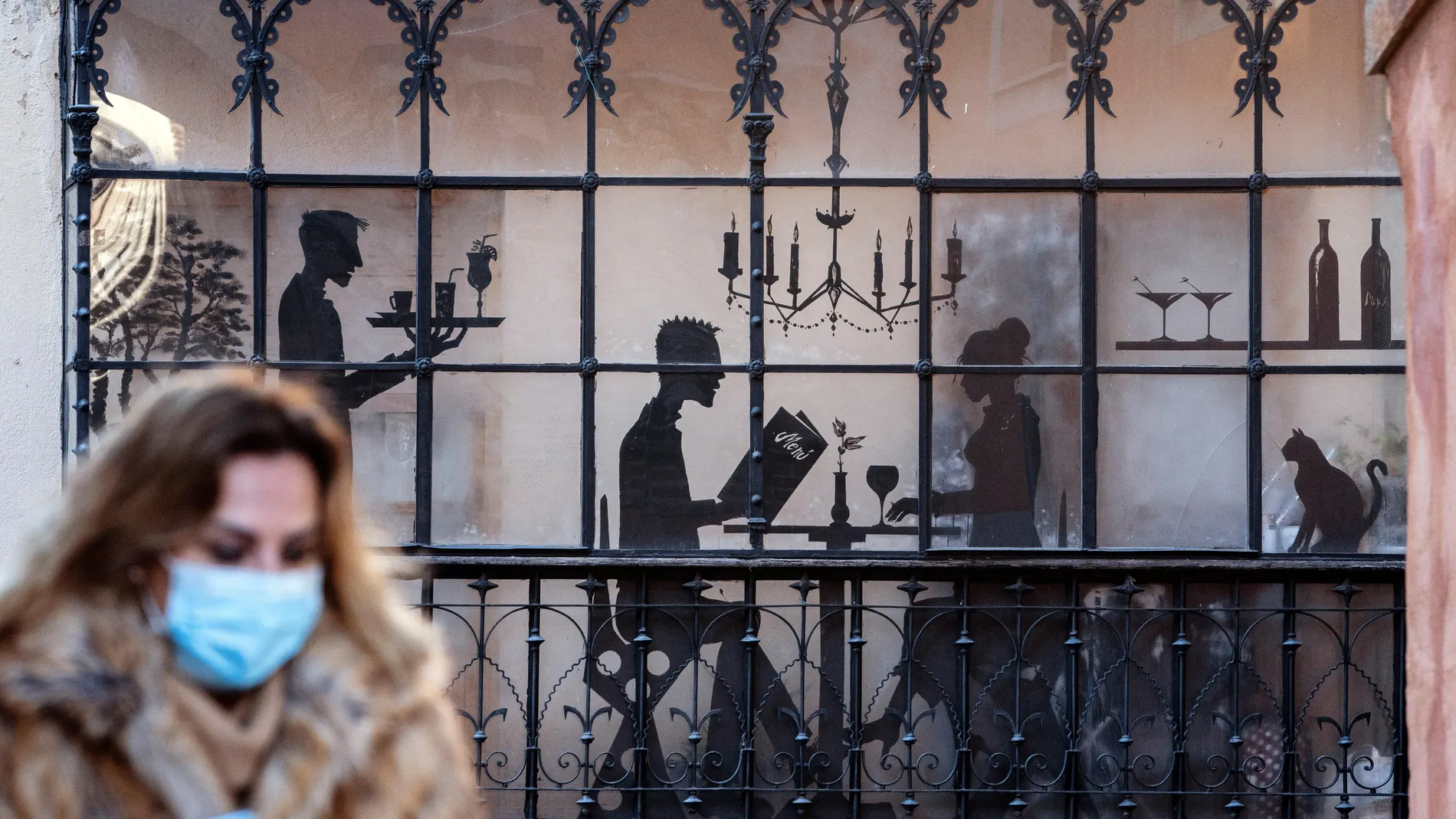 Una mujer con mascarilla pasa por delante de la fachada de un restaurante decorado con siluetas negras de camareros y comensales, este lunes en Ávila. La hostelería se prepara para afrontar la Nochevieja pendiente de posibles cambios en las restricciones para contener la pandemia del Covid-19. EFE/ Raúl Sanchidrián