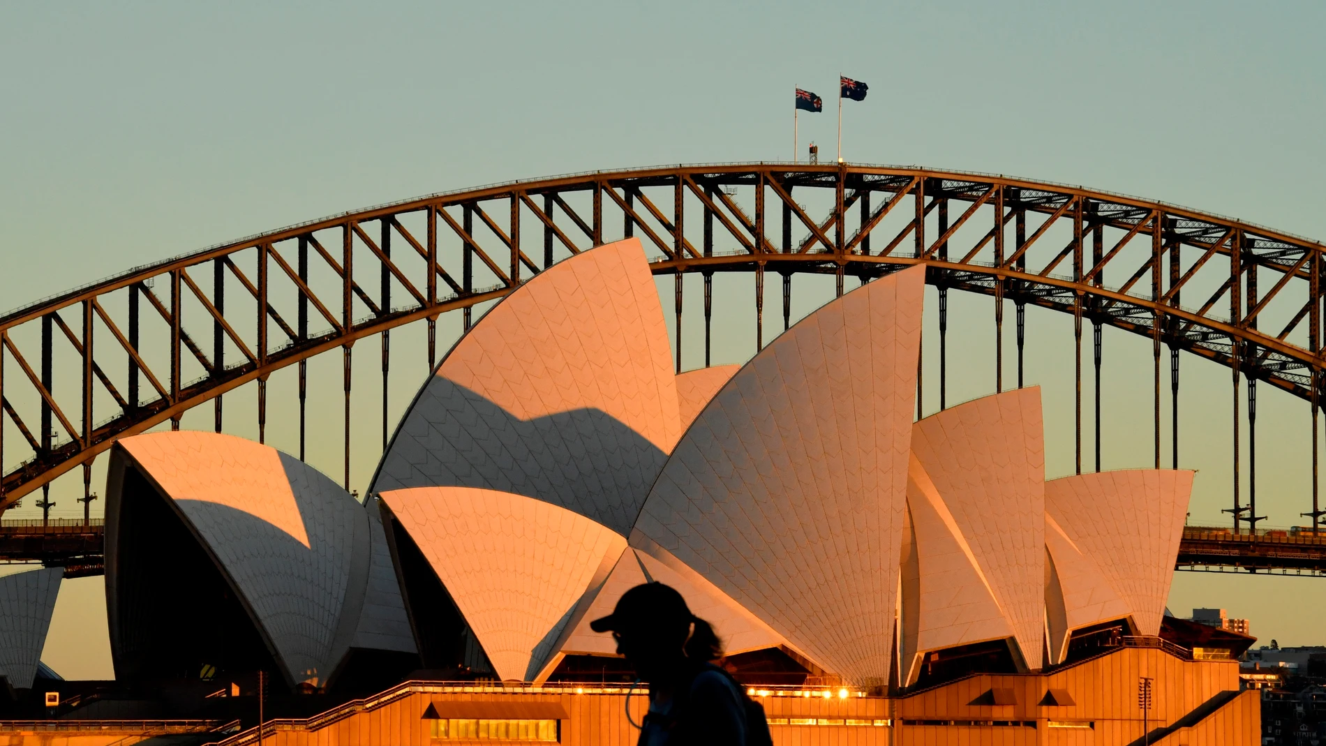 Una mujer camina junto a la Ópera de Sidney durante el amanecer. Unos 300 soldados australianos se unirán, a partir del lunes, a las tareas de vigilancia de la Policía para garantizar el cumplimiento de las restricciones impuestas por un brote con la variante delta de la covid-19