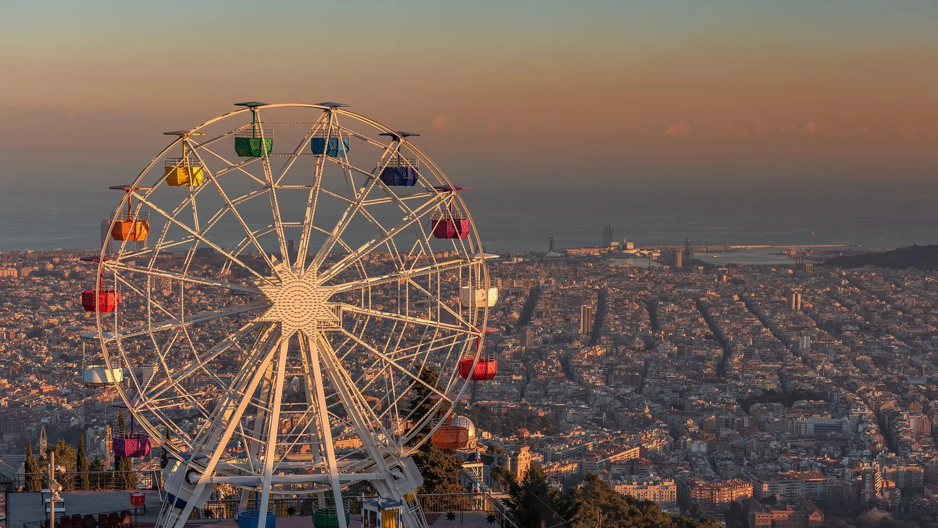En primer plano se ve una noria blanca con cabinas de colores. Detrás en un plano más bajo se ve la ciudad de Barcelona, y al fondo se ve el cielo azul con una capa naranja y gris de contaminación