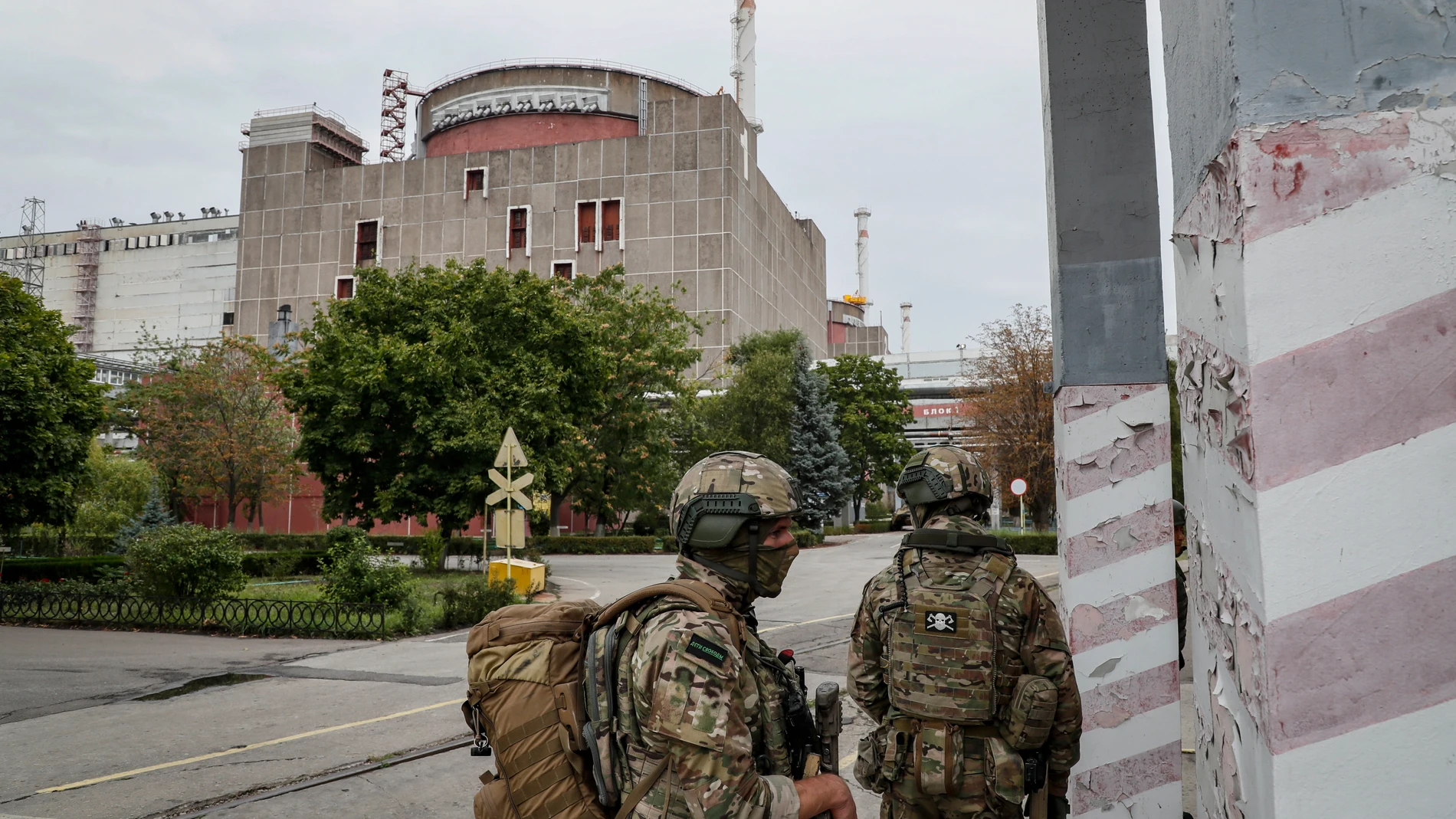 Al fondo, un edificio gris con un cilindro rojo en medio. En primer plano, dos soldados armados