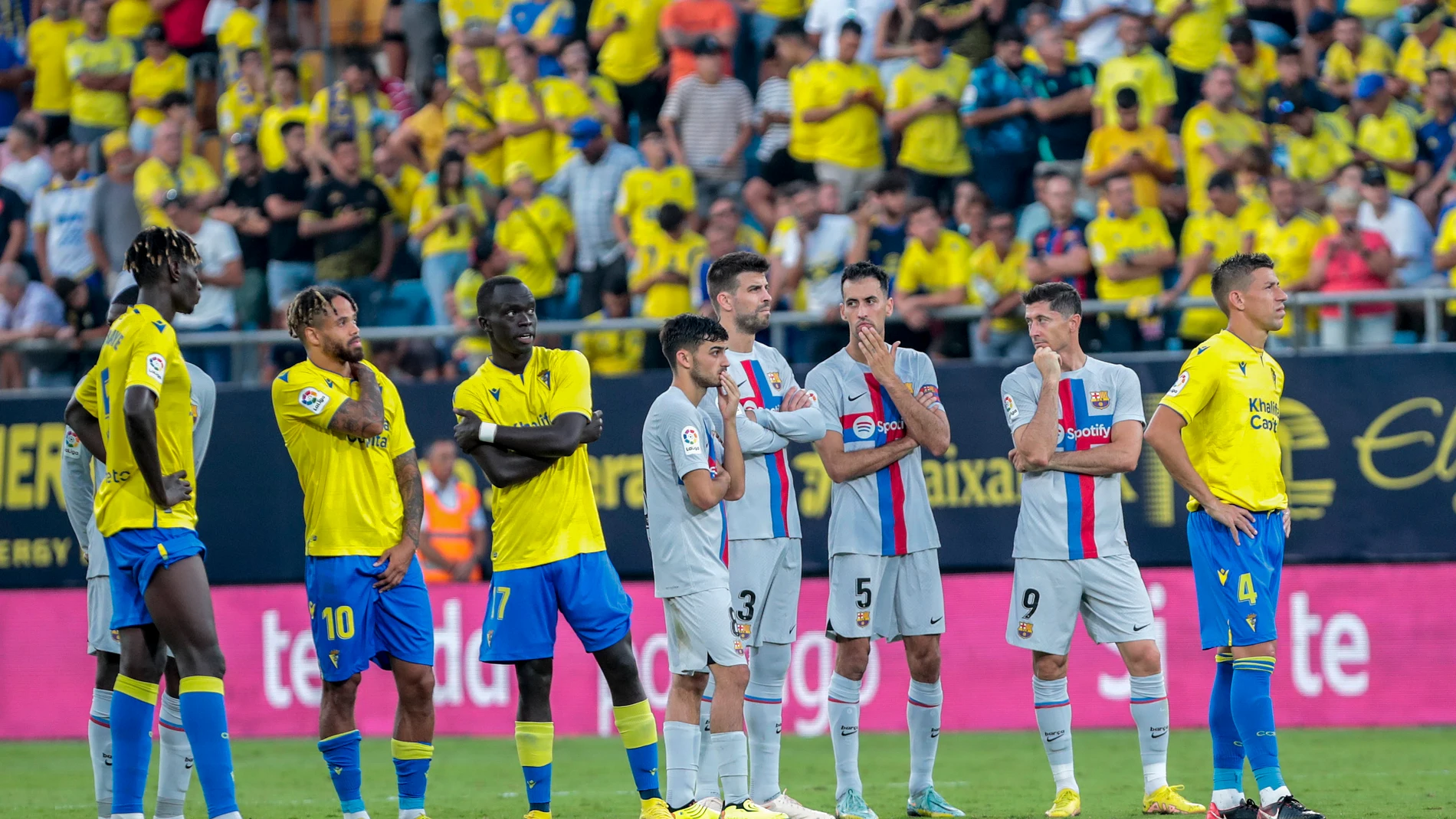 Los jugadores del Barcelona y el Cádiz esperan en el césped, preocupados por lo que sucede en la grada