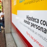 Cartel de hipoteca en una entidad bancaria con una persona pidiendo limosna al fondo.