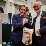 El alcalde de Madrid, José Luis Martínez-Almeida y Luís Cueto le entrega un banderín de su nueva formación política “Recupera Madrid”, durante el pleno del Ayuntamiento de Madrid.