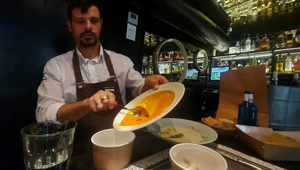 El camarero del Mercado de la Reina, Gabriel Biglia, echa en el táper de Envapro el salmorejo sobrante: "Normalmente lo haríamos en la cocina", cuenta a este reportero. A la derecha, podemos ver el envase biodegradable "más grande que usamos" y la bolsa de papel que dan a los comensales.
