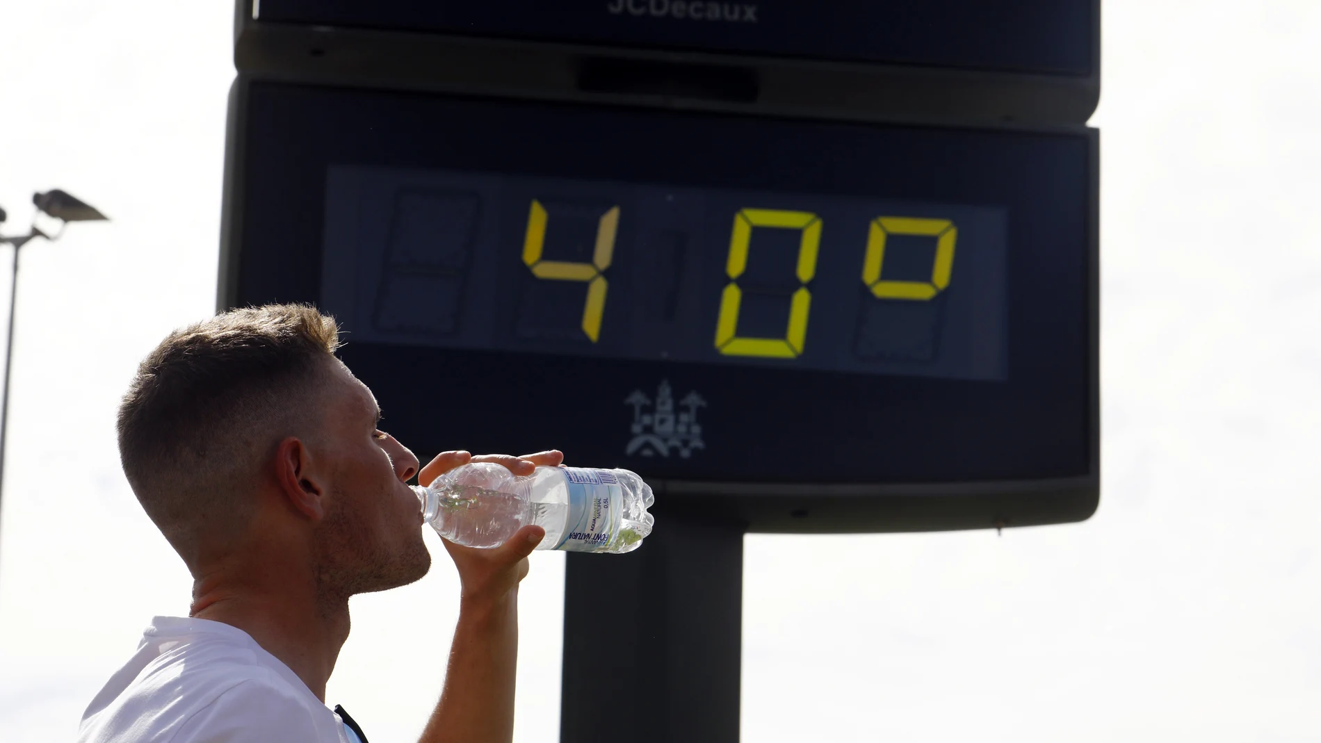 Un joven bebe agua junto a un termómetro de calle que marca 40 grados