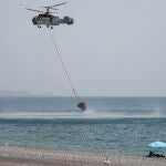 Un par de bañistas ven desde la playa un helicóptero que se llena del agua del mar durante un incendio forestal, cerca de la aldea de Gennadi, en la isla griega de Rodas, al sureste de Grecia, hoy jueves 27 de julio. Rodas es una isla muy turística donde han sido desalojadas 20.000 personas por los incendios