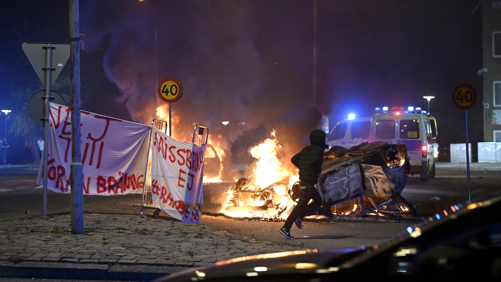FILE - A person runs past a burning car and a banner reading, "No to Quran burning. Don't abuse freedom of expression" placed on the roundabout, at Ramel väg, in Malmo, Sweden, early Monday Sept. 4, 2023. The targeting of Swedish citizens in an attack in Brussels on the night of Monday, Oct. 16, 2023 has shocked the Scandinavian country, yet the government had been warning for months that Swedes were at greater risk since a recent string of public desecrations of the Quran, Islam's holy book,...
