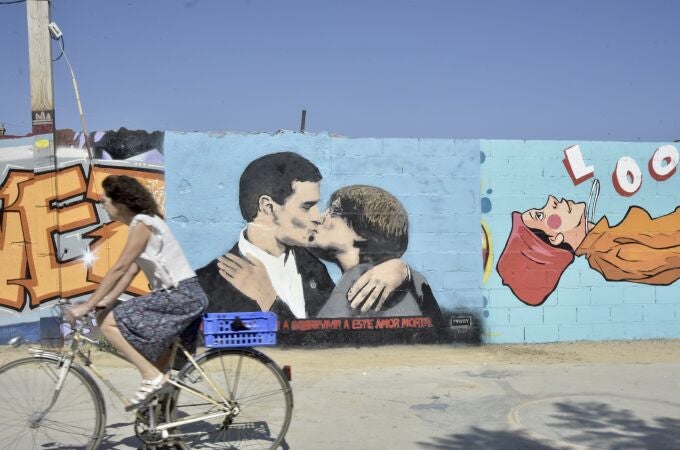 Un ciclista pasa al lado del mural del presidente español y candidato del PSOE a la reelección Pedro Sánchez y al expresidente de la Generalitat y eurodiputado de Junts, Carles Puigdemont, dándose un beso en el mural del artista urbano TVBoy, en la plaza de las Glòries, en Barcelona, 