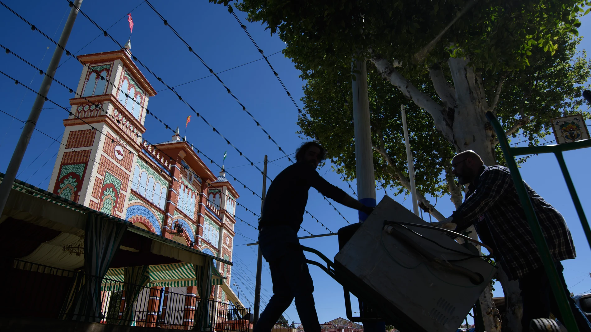 SEVILLA, 09/04/2024.- Dos hombres transportan este martes una nevera para una de las casetas del recinto de la Feria de Abril de Sevilla, que arranca el próximo fin de semana. EFE/ Raúl Caro