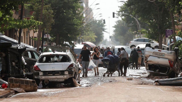 VALENCIA, 30/10/2024.- Unas personas caminan por una calle cubierta de lodo y llena de coches amontonados tras las intensas lluvias por la fuerte DANA hoy en Valencia