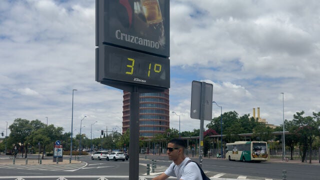 Un hombre montando en bicicleta pasa junto a un termómetro que marca 31 grados en Sevilla