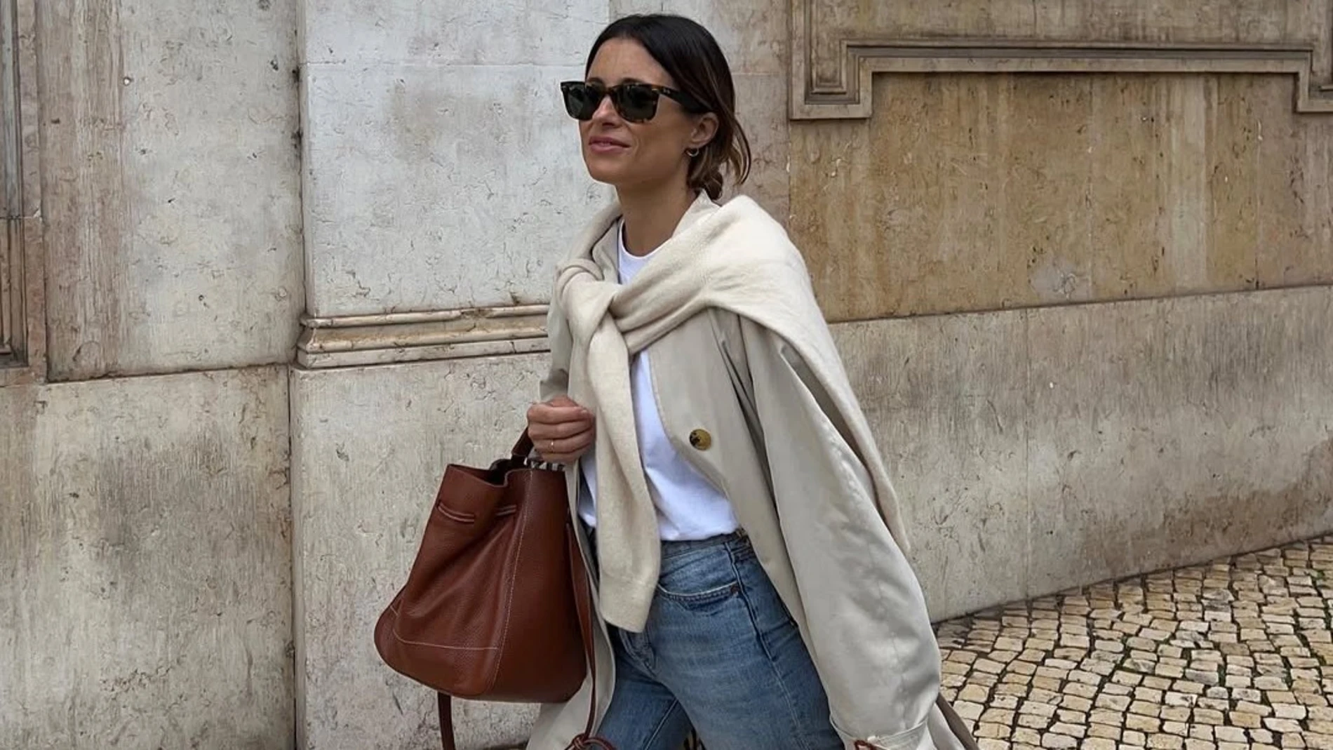 Influencer con gabardina beige larga, vaqueros azules rectos, camiseta blanca básica y jersey claro sobre los hombros. Completa el look con bolso marrón de piel y gafas de sol oscuras.