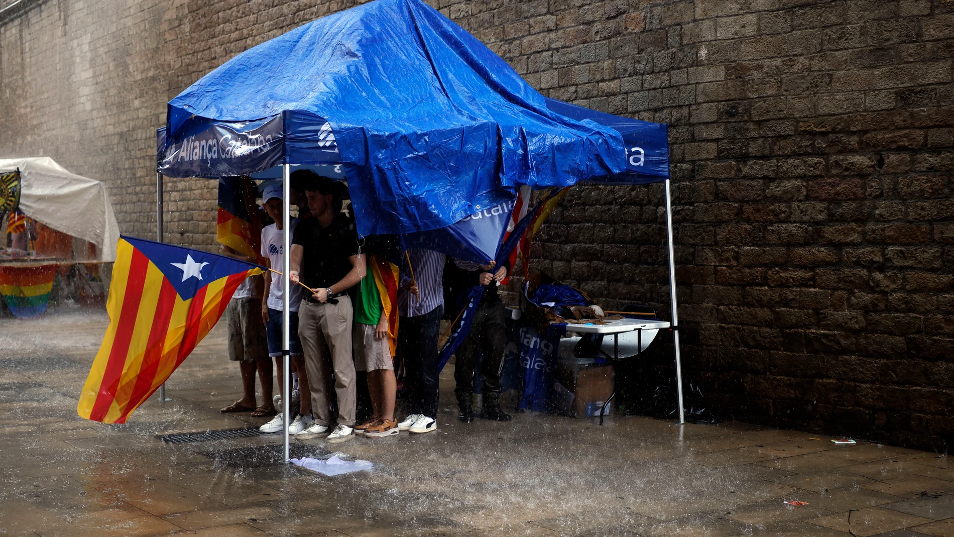 GRAFCAT1933. BARCELONA, 11/09/2025.- Varias personas se protegen bajo una carpa de Aliança Catalana de la intensa lluvia que este jueves ha caído en Barcelona y que ha obligado a suspender algunos de los actos previstos para la celebración de la Diada. EFE/Alberto Estévez
